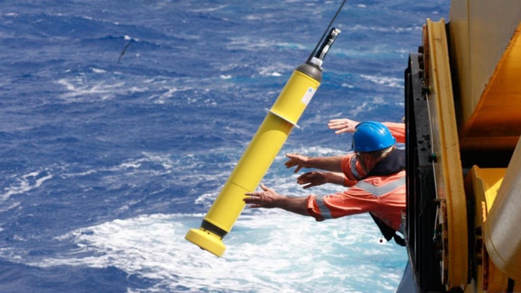 Two men outstretched over the side of the ship in orange life jackets and long sleeve orsnge tee shirts throw a human-sized yellow instrument with a block rod at the top filled with sensors into the whitewater and the ocean filling the picture