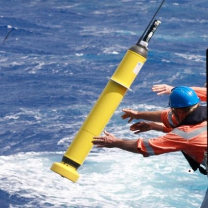 Two men outstretched over the side of the ship in orange life jackets and long sleeve orsnge tee shirts throw a human-sized yellow instrument with a block rod at the top filled with sensors into the whitewater and the ocean filling the picture