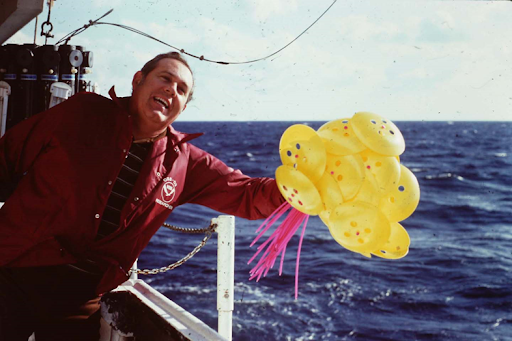 A male scientist dressed in a red full-body jumpsuit releases a handful of yellow scientific instruments, called current drifters, off the side of a ship