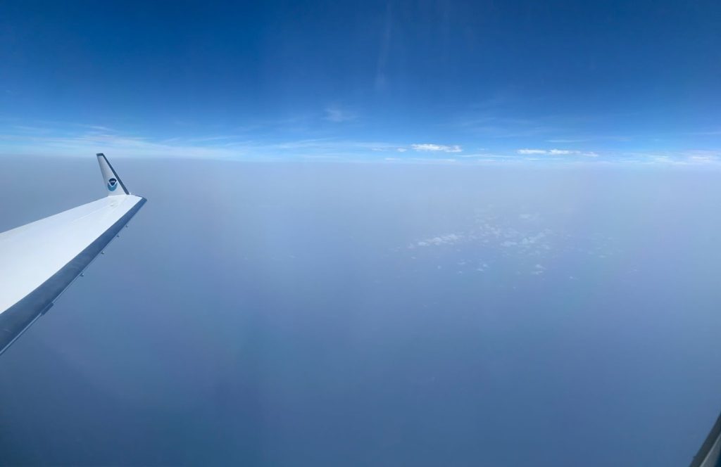 A view of the Saharan Air Layer over the Atlantic Ocean from the port side of a NOAA G-IV Hurricane Hunter aircraft. Blue sky above with a greyish white haze tinting the ocean below. The airplane wing extends from the left side of the frame. Expanding the scope of study and exploring tropical waves at their source helps noaa forecast models predict future track and intensity.