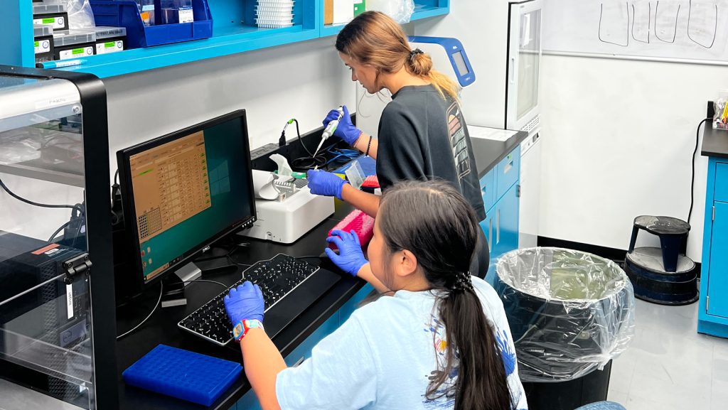 Two interns work at a black counter in a lab, diving into 'Omics analyses. Intern Lorelei Ing sitting in a light blue shirt with a long pony tail sits at a computer and enters data at the screen in front of her while wearing blue gloves. Taylor Gill is to her right with brunette hair in a bun and a black shirt, wearing blue gloves as she holds a pipette in right hand. In front of her is a white, cubed NanoDrop device.