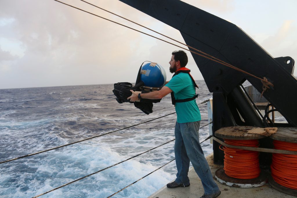 Researcher Shaun Dolk holds a round ocean current drifter off the side of a ship before tossing it into the ocean to gather data.