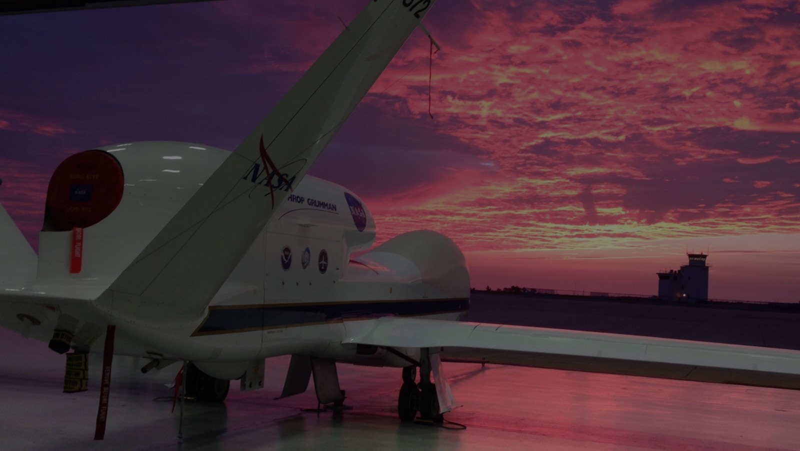 Photo of the NASA Global Hawk in the hangar going out into a bright pink sunset. Photo Credit, NOAA.