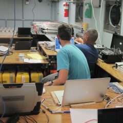 An AOML scientist (right) explains to one of the university students participating in the cruise how data is acoustically downloaded from one of the NOAA subsurface moored instruments. Image credit: NOAA