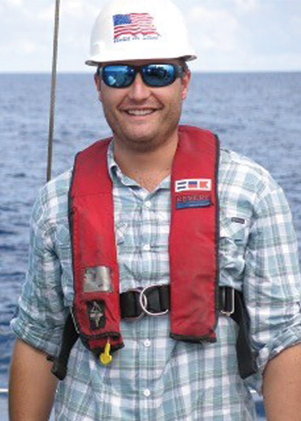 Retrato en color de un Michael Studivan sonriente con casco blanco, chaleco salvavidas autoinflable rojo con tirador amarillo, gafas de sol de espejo azules con montura blanca y camisa de cuadros azul verdoso, gris y blanco con las mangas remangadas.