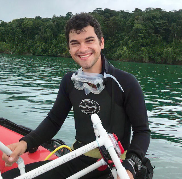 Fotografía en color de Ben Chomitz sentado en el borde de un barco con equipo de buceo y equipo de muestreo de tubos de PVC.