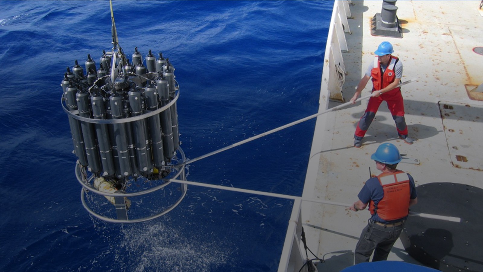 Two NOAA scientists deploy a large CTD into the ocean.