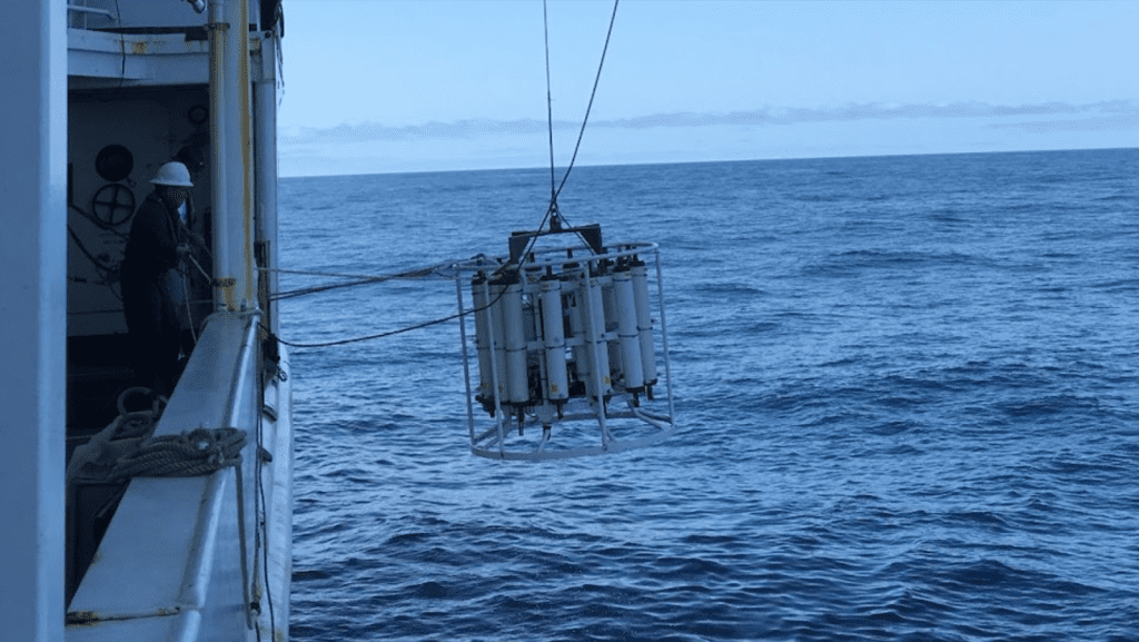 A large white CTD being deployed into the ocean off the side of a side.