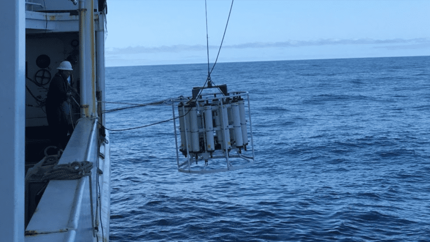 A large white CTD being deployed into the ocean off the side of a side.