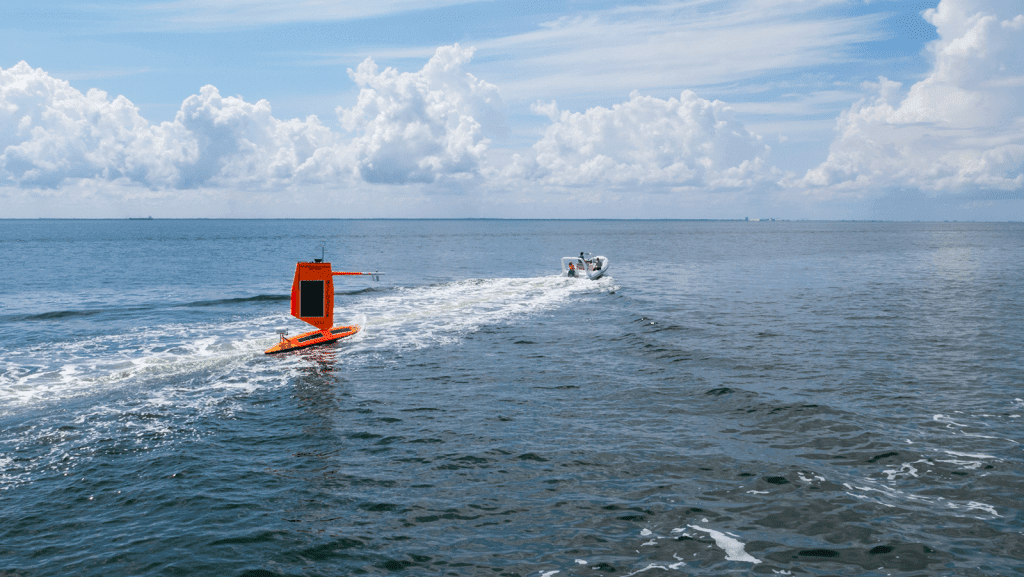 A large orange saildrone is in the water being towed by a small boat.