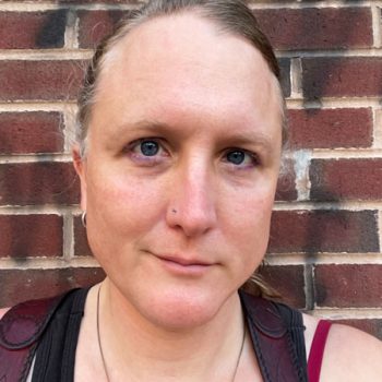 Portrait of Thia Griffin-Elliott, a caucasian trans/nonbinary femme person with nose piercing a half smile blue eyes and smudged makeup wearing a black shirt and red shoulder harness in front of a brick wall