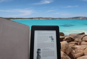 A black kindle is being held with a body of blue water in the background.