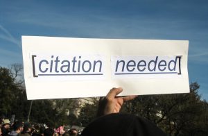 A person's hand against a blue sky holding up a sign that reads "citation needed".