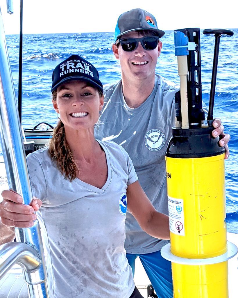 Jenn McWhorter and another researcher with BGC Argo, Image of a caucasion man and a caucasian woman standing and looking into the camera, both in grey shirts with baseball hats on the port side of a small center console boat looking forward, holding an oblong cylindrical Instrument with a white colar and a black top with sensors and an antennae protruding from the top, balanced on the edge of the boat