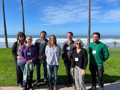 Seven scientists wearing name tags pose for a photo with a green lawn, palm trees, and the Pacific Ocean in the background.