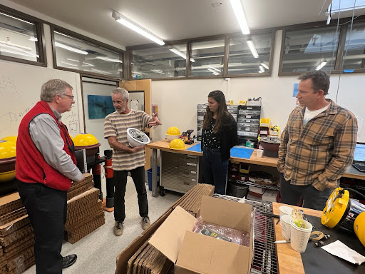 Four scientists receive a tour of a laboratory where yellow and white scientific instruments are on display.