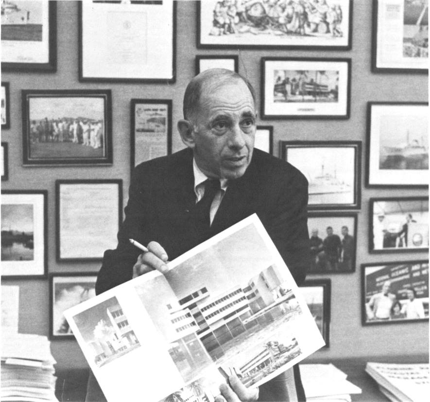 A Black and White image from AOML's Archive of AOML's first director Dr. Harris B. Stewart Jr. in his office holding a magazine with a two-page photo of AOML's building.