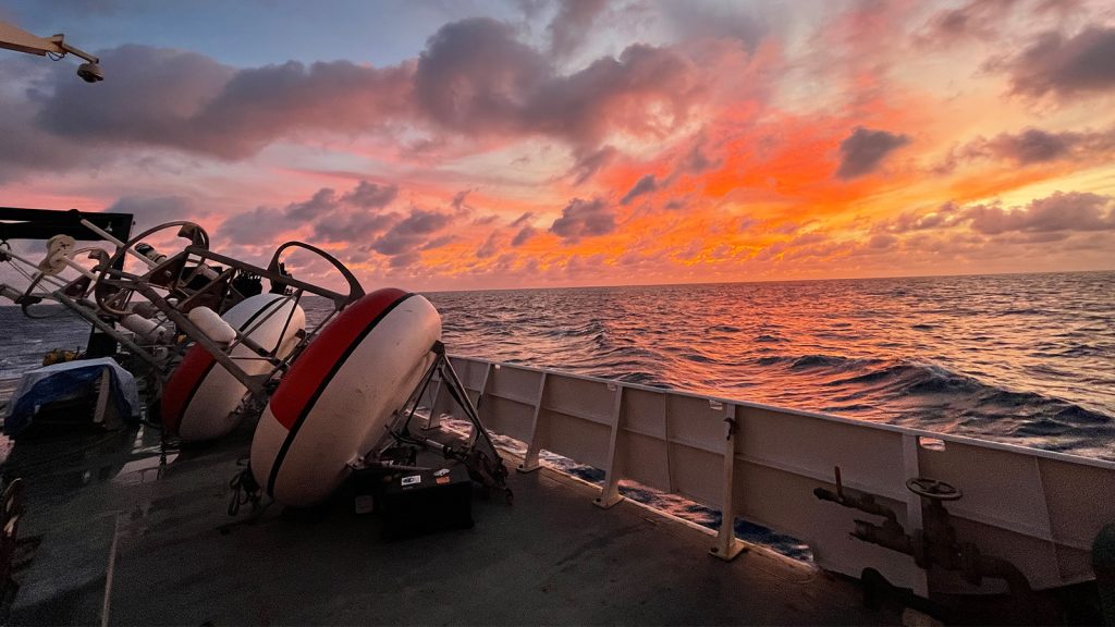 Varias boyas PIRATA tumbadas a bordo del buque Ronald H. Brown de la NOAA frente a un cielo rosa, amarillo y naranja durante el crucero PIRATA Northeast Extension (2022).