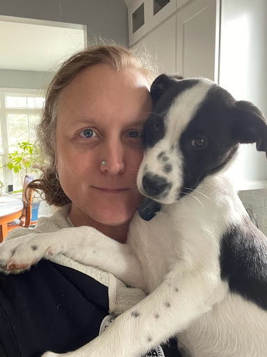 Color photograph of Thia Griffin-Elliott, a caucasian trans femme/nonbinary individual, holding a black and white 3-month old puppy.