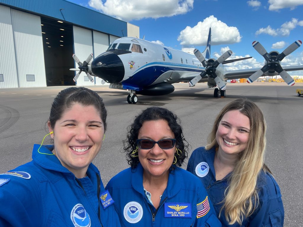 Color Photograph of 2023 Hurricane Field Program Deputy Director Heather Holbach (left), Hurricane Research Division Deputy Director Shirley Murillo (center), and former AOML researcher Samantha Camposano (right) in front of p-3 orion hurricane hunter,  after their research mission into Hurricane Fiona