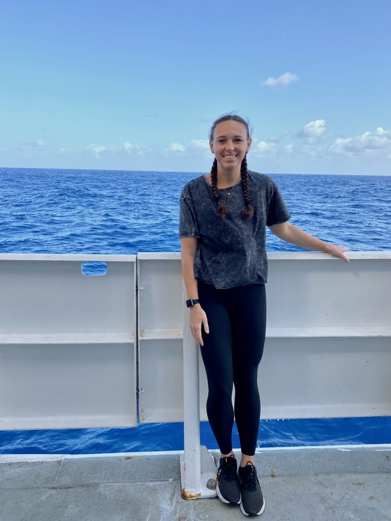 La Dra. Katelyn Schockman, científica del AOML/CIMAS, de pie y sonriendo junto a la barandilla de un barco.Photo Credit: NOAA/AOML