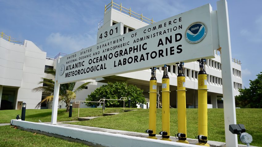 A sign with the words "Atlantic Oceanographic and Meteorological Laboratory" stands in from of a building and there are yellow scientific instruments being displayed in front of the sign.