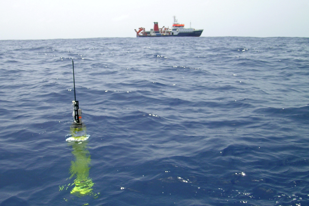 Flotador biogeoquímico amarillo en el océano, en primer plano, con un barco a lo lejos. Este flotador está transmitiendo datos y recibiendo órdenes antes de realizar otra inmersión para recoger datos.