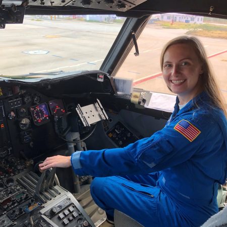AOML/CIMAS communication intern Holly Stahl in a blue NOAA flight suit sitting in the cockpit of a NOAA P-3 Hurricane Hunter aircraft.