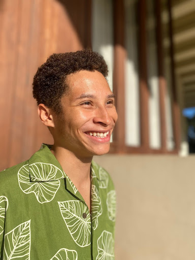 Akili Ligons smiling and wearing a green, leaf patterned button down and looking off into the distance 
