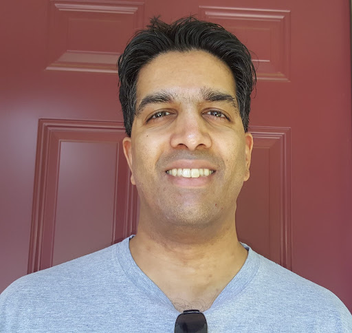 Ashvin smiles at the camera with brown hair and a silver tee shirt in front of a red door. 