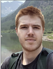 Color selfie of Nicholas MacKnight in front of a lake in the mountains, wearing a backpack