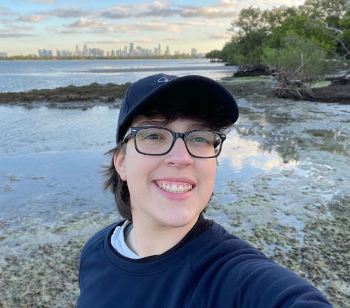 ALT: Nina Castro Alves smiling with a skyline behind her, wearing a hat and glasses 
