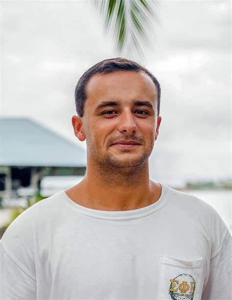 Alt: Chris Malanuk grimaces at the camera in front of a dock with a green roof. He has a white shirt and a cloudy sky with a single palm dangling above him. 