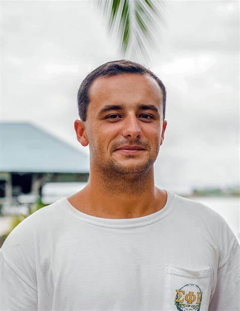 Alt: Chris Malanuk grimaces at the camera in front of a dock with a green roof. He has a white shirt and a cloudy sky with a single palm dangling above him.