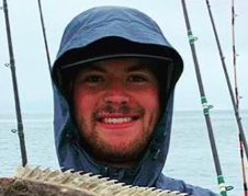 Sterling smiles at the camera with a hat and gray rain jacket on to shield from expected rain. Fishing poles and blue ocean are behind him. 