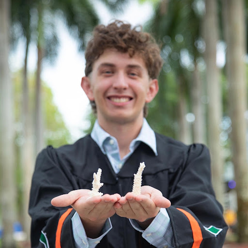 Zachary Zagon poses for the camera in a blue collared shirt with his robes on as he graduates. He holds two pieces of coral in his hands, completely white. The background is faded though we can see an aisle of palm trees on both sides of him.