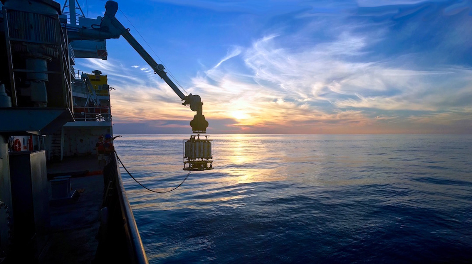 Sunset photo: The sun dips to the surface of the glassy blue ocean with few white clusters of clouds. The ship is taken fromthe side of a vessel as we can see the boat's hull along the left edge of the photo, a niskin bottle sampler being lowered into the ocean over the side almost next to the sun.