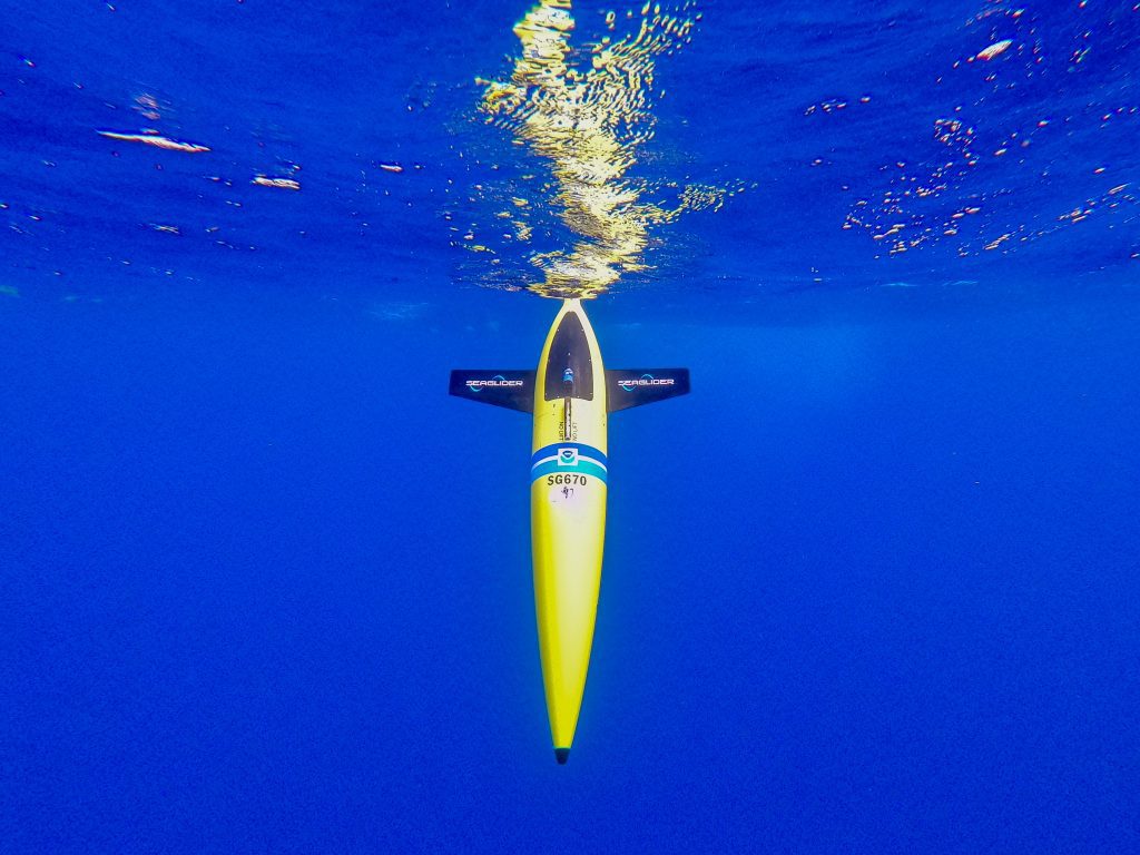 Foto submarina del océano azul con un planeador amarillo sentado verticalmente en el agua, preparado para realizar la inmersión programada para recoger datos oceánicos.