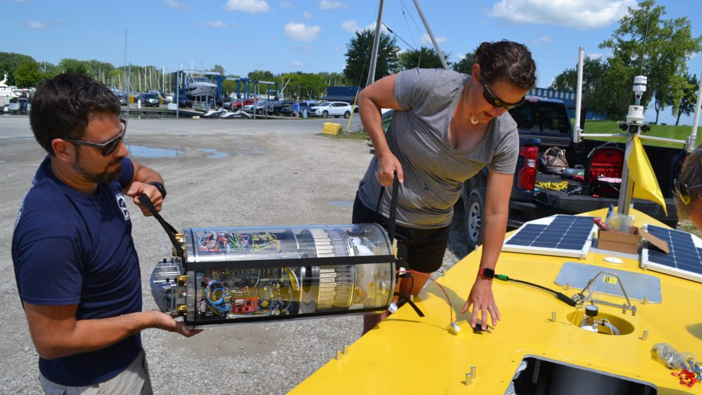 A man in a bue shirt and sunglasses (left) and a woman in a gray tee with dark sunglasses hoist the massive 3G ESP into the center of the SeaTrac, a yellow and large horizontal board taking up mainly the bottom right corner of the photo. The people ar in the center and between them is the 3G ESP, which we can tell is a massive and seeminlgy heavy barrel shaped glass container of tubes and wires neatly compact within it. The yellow SeaTrac has a well-fixed hole in the middle of it that we can see the 3G ESP is being loaded into carefully. They do this in a boat yard while the SeaTrac is still just on a trailer attached to a black truck (whcih we can see behind thetwo people carrying the 3G ESP). 