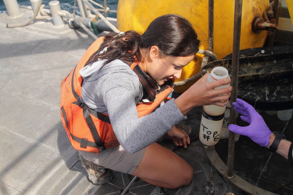 Woman with brown hair, gray shirt, and orange life vest squats down on a ship and holds up a white bottle.
