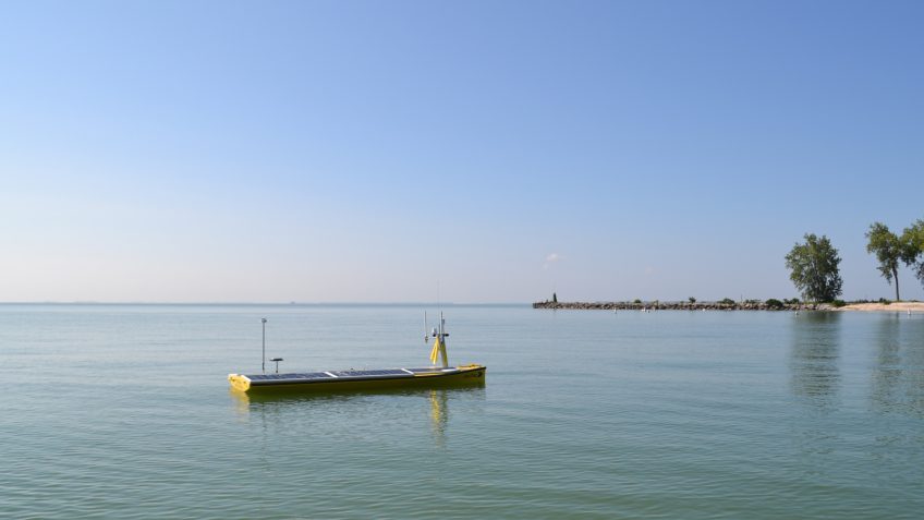 On a beautiful calm day, the SeaTrac, a thin yellow vessel roughly the shape of a stand up paddleboard, glides aong the glassy waters of the Great Lakes, easily navigating the shallows with trees and jetties of rocks beyond it and a sea bordering the skies somewhere that we can barely define.