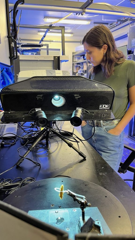 Kenzi Cooke scans a coral fragment (a small piece maybe an inch tall) held in place by a ring with a beam of light shining on it from the large black projector taking up the center of the photo.