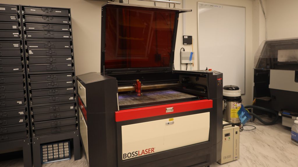 AMDL_Laser-Cutter The cubed laser cutter with the top open to displace the red metal laser that moves along a silver metal bar on the interior