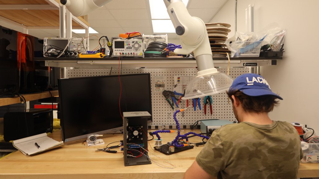 Nash in a blue hat works at the soldering station in the AMDL on a wooden counter with wires and cables and blue wired holders with metal clamps holding a red circuit board fixed as he works