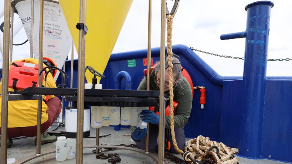 Emma Graves kneels next to the yellow cone-shaped sediment trap with a black   rounded disc at the bottom, adding a series of white plastic cups to the underside of the black disc in a rotating, circular array. 