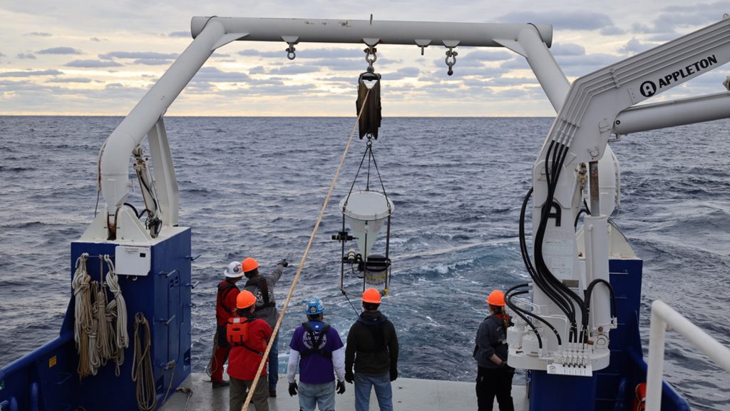 Six people in jeans and red and black rain jackets and orange helmets stand at the stern of a research vessel beneath a massive white crane with a white cone-shaped sediment trap swaying before them from a rope as its carried from the depths of the Gulf of Mexico with only the ripples of an uneven yet vast open ocean beyond where they all stand. 