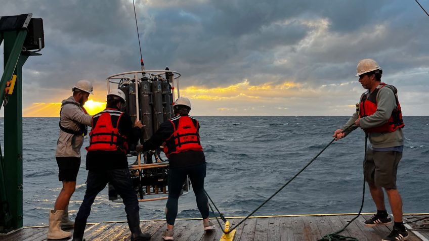 Three scientists and ocean engineers in orange lifejackets keep the CTD steady as it leans with the boat in rough seas while the other scientist cleats the CTD with a long rope, we see big waves and the sunset below a sky of cloud