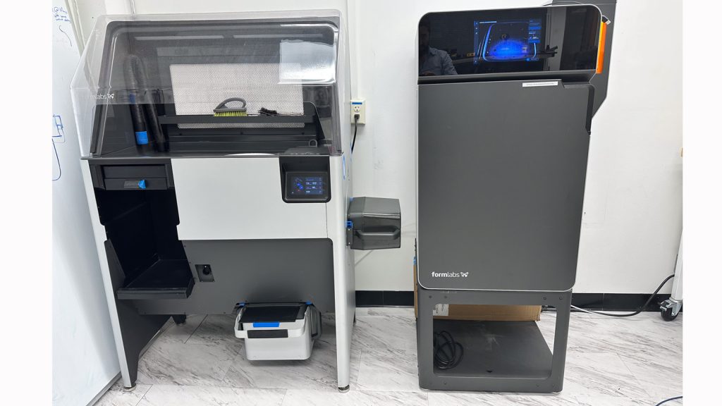 THe Fuse tall rectangular black printer on the right with the dust cleaning station (also metallic grey) on the left with a white background tiled floor