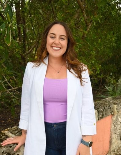 Bailey Morris in a white coat and a purple shirt with green trees behind her