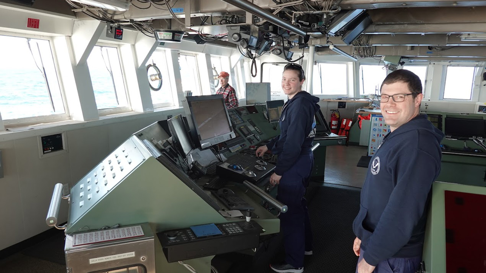 NOAA CORPS OFFICER ANNA GASKILL AT THE HELM of a massive marine vessel in the cockpit with a white interior and massive windows aligned the walls peering out onto the blue ocean outside, she wears a blue uniform and stands smiling with a coworker and officer to her right with dark hair, glasses and the same blue outfit